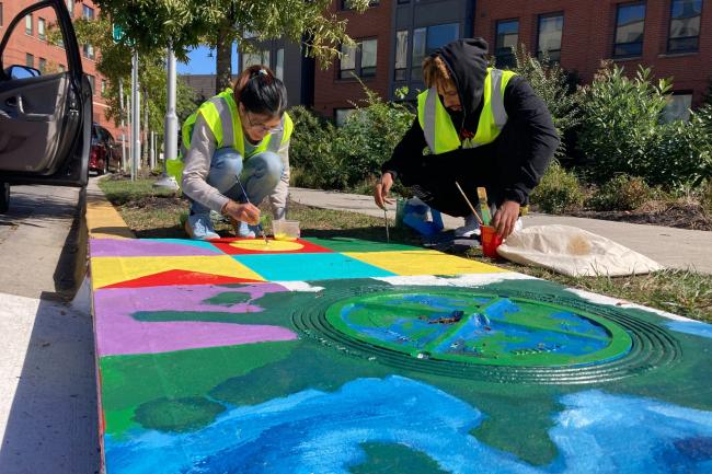 Carla Duran and Beniyam Berhanu, UMD student artists, paint a storm drain along Guilford Drive on Oct. 10, 2022. (Rachel McCrea/For the Diamondback)